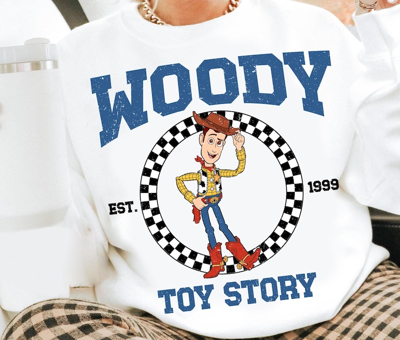 Puede incluir: Sudadera blanca con un gr&aacute;fico de Woody de Toy Story. El dise&ntilde;o incluye la palabra "WOODY" en letras azules en bloque sobre una imagen circular del personaje. Las palabras "EST. 1999" y "TOY STORY" tambi&eacute;n est&aacute;n impresas en la sudadera.
