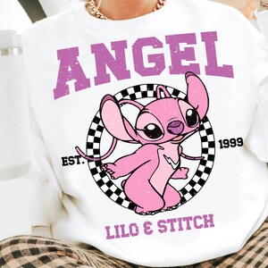 Puede incluir: Sudadera blanca con un dise&ntilde;o de personaje de dibujos animados rosa. La palabra "ANGEL" est&aacute; impresa en p&uacute;rpura sobre el personaje, con "LILO & STITCH" debajo. El personaje est&aacute; dentro de un c&iacute;rculo a cuadros en blanco y negro.