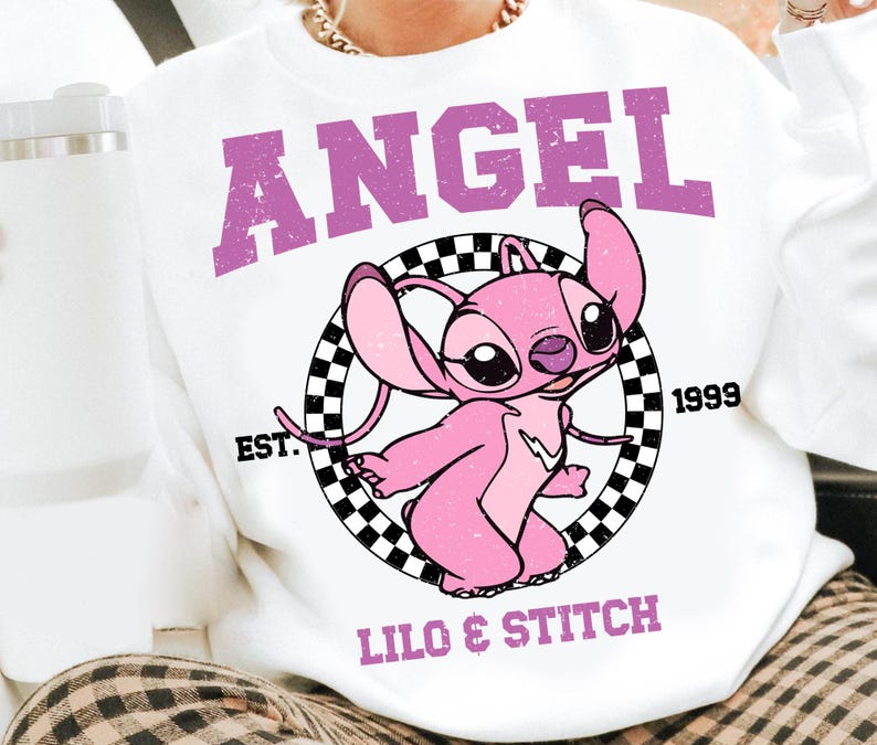 Puede incluir: Sudadera blanca con un dise&ntilde;o de personaje de dibujos animados rosa. La palabra "ANGEL" est&aacute; impresa en p&uacute;rpura sobre el personaje, con "LILO & STITCH" debajo. El personaje est&aacute; dentro de un c&iacute;rculo a cuadros en blanco y negro.