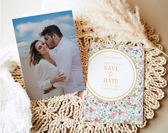 May include: A wedding save-the-date card with floral patterns and a photo of a couple kissing on a beach. The card has gold text that reads "Save the Date" and the date April 26, 2024. The photo is on the left, and the card is on a woven mat.