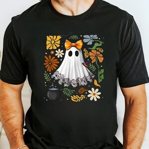 May include: Black t-shirt featuring a white ghost with an orange bow, surrounded by colorful flowers and leaves. The ghost has a lace trim and is wearing a witch hat. A cauldron is at the bottom left of the design. Halloween theme.