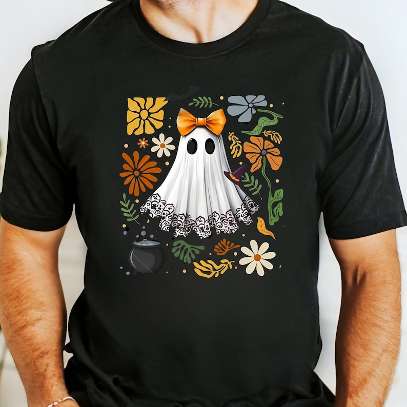 May include: Black t-shirt featuring a white ghost with an orange bow, surrounded by colorful flowers and leaves. The ghost has a lace trim and is wearing a witch hat. A cauldron is at the bottom left of the design. Halloween theme.