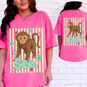 May include: A bright pink t-shirt featuring a brown monkey graphic with the word "See" in turquoise letters. The design includes yellow and white striped background, turquoise flowers, and pink stars. The back of the shirt has a similar design with the word "Do."