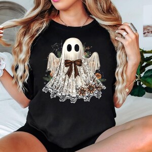 May include: Black t-shirt featuring a ghost design. The ghost is white with black eyes and a brown bow. The ghost is wearing a lace shawl and is surrounded by flowers and a spider web. The shirt is a crew neck.