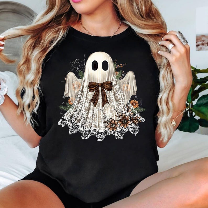 May include: Black t-shirt featuring a ghost design. The ghost is white with black eyes and a brown bow. The ghost is wearing a lace shawl and is surrounded by flowers and a spider web. The shirt is a crew neck.
