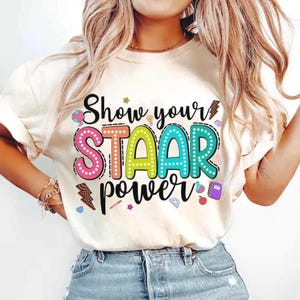 Peut inclure: T-shirt crème avec l'inscription "Show your STAAR power" en lettres à bulles colorées. Le t-shirt présente un design ludique avec des éclairs, des crayons et un cahier.