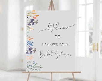 May include: Welcome to Harlowe James Bridal Shower sign with a watercolor floral border in shades of blue, orange, and purple. The sign is on an easel.