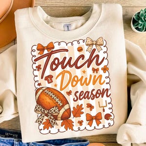 May include: Cream-colored sweatshirt with a football-themed graphic. The design includes a football, bows, leaves, and the words "Touch Down season" in brown and orange. The sweatshirt is a casual, fall-themed apparel item.
