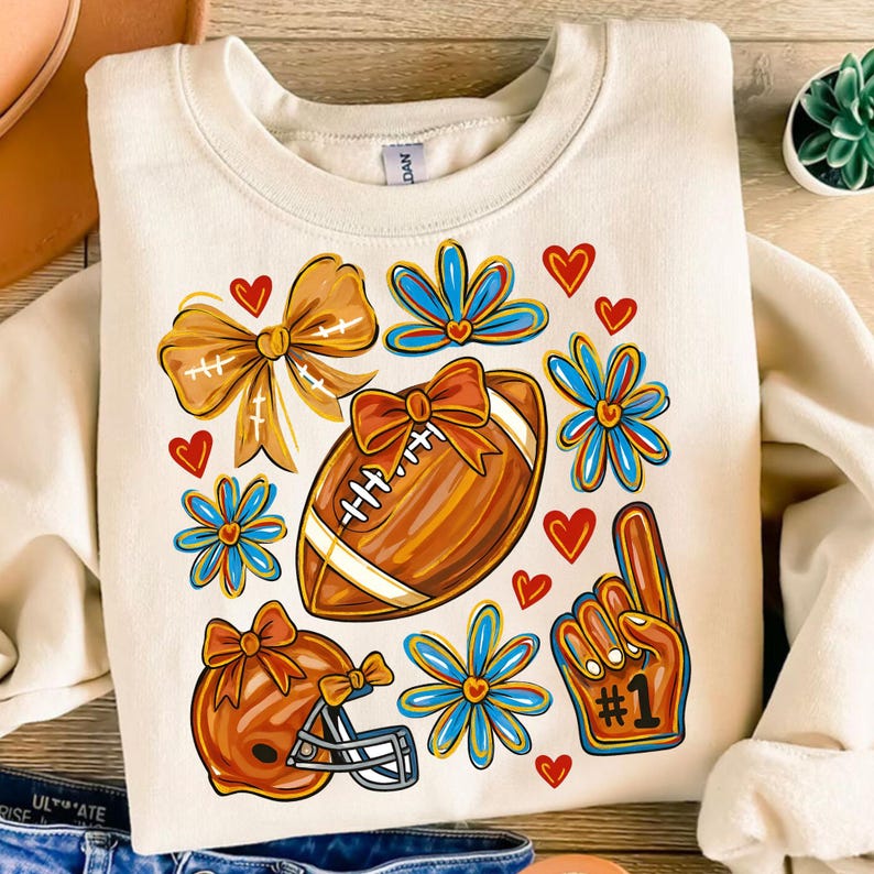 May include: Cream-colored sweatshirt featuring a football, helmet, and #1 foam finger graphic. The design includes orange bows, blue flowers, and red hearts. A fun and festive design for football fans.