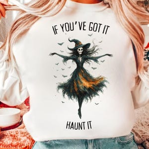 May include: White sweatshirt with a Halloween-themed graphic. The graphic features a skeleton witch with a black dress and hat, surrounded by bats. The text "IF YOU'VE GOT IT HAUNT IT" is printed above and below the witch.