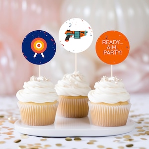 May include: Three cupcakes with white frosting and gold sprinkles. Each cupcake has a round paper topper with a different design. One topper is blue with a target, one is white with a cartoon laser gun, and one is orange with the text "READY... AIM... PARTY!"