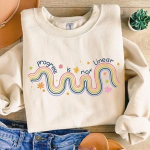 May include: Cream-colored sweatshirt with the text "progress is not linear" in a colorful, rainbow-like design. The design includes flowers and stars. The sweatshirt is folded, with a pair of blue jeans and sandals visible in the background.