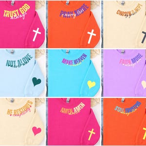 May include: A collection of colorful sweatshirts with inspirational phrases. Each sweatshirt features a different color, such as pink, orange, and blue, with text like "Trust God" and "Pray More." A cross or heart graphic is on the sleeve.