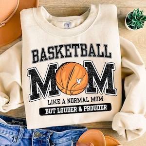 May include: Cream-colored sweatshirt with the text "BASKETBALL MOM" in black block letters. A basketball graphic is in the center of the word "MOM". Below the text is "LIKE A NORMAL MOM BUT LOUDER & PROUDER" in black.