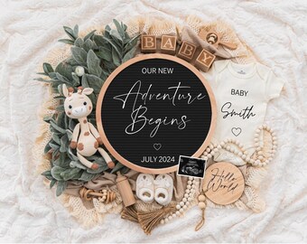 May include: A black letter board with the text "Our New Adventure Begins" and "July 2024".  The board is surrounded by a variety of baby items, including a stuffed giraffe, a baby onesie, a wooden block that says "Baby", and a wooden sign that says "Hello World".