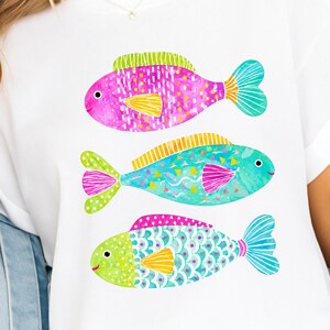 Op de afbeelding: Wit T-shirt met drie kleurrijke visillustraties. De vissen zijn geschilderd in roze, turquoise en groen, met gele vinnen. Elke vis heeft een uniek patroon, wat zorgt voor een speels en artistiek ontwerp.