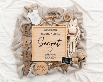 May include: A wooden sign with the text "We've been keeping a little secret. Arriving July 2024."  The sign is surrounded by a beige knitted blanket, wooden letters spelling "BABY", a wooden toy bunny, a wooden rattle, and a small wooden sign with the text "Hello World".