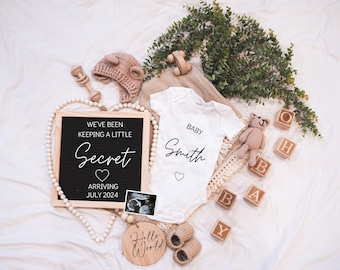 May include: A black letter board with the text "We've been keeping a little secret arriving July 2024" and a heart. A white onesie with the text "Baby Smith" and a heart. A wooden sign with the text "Hello World". A teddy bear, a wooden toy, and a baby hat.