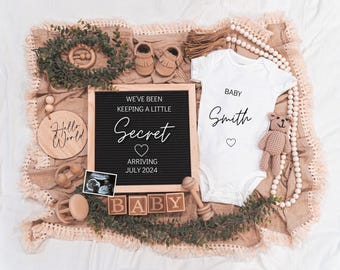 May include: A black letter board with the text "We've been keeping a little secret arriving July 2024" and a white onesie with the text "Baby Smith" on it. The onesie is surrounded by wooden toys, a knitted teddy bear, and a wooden sign that says "Hello World".