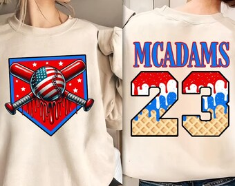 May include: Cream-colored sweatshirt featuring a baseball-themed graphic. The front displays a baseball with an American flag design, crossed bats, and a red, white, and blue color scheme. The back has the name "MCADAMS" and the number "23" with a waffle pattern.
