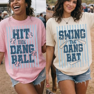 Op de afbeelding: Twee vrouwen dragen honkbal-thema T-shirts. Eén shirt is lichtroze met de tekst "HIT THE DANG BALL" in blauw, samen met honkbalafbeeldingen. Het andere shirt is crèmekleurig met "SWING THE DANG BAT" in blauw.