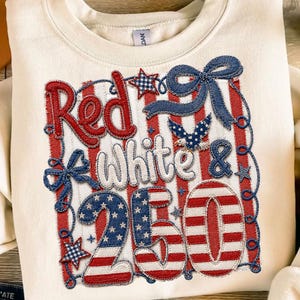 May include: Cream-coloured sweatshirt with a patriotic design. The design features the words "Red White & 250" in red, white, and blue, with stars and stripes. A blue bow and an eagle are also included in the design.