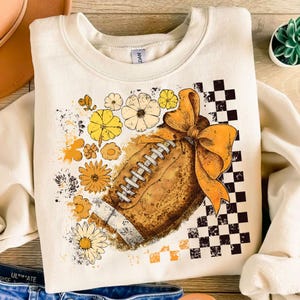 May include: Cream-colored sweatshirt featuring a football design with a large orange bow and floral accents in yellow and white. A black and white checkered pattern is on the right side of the design.