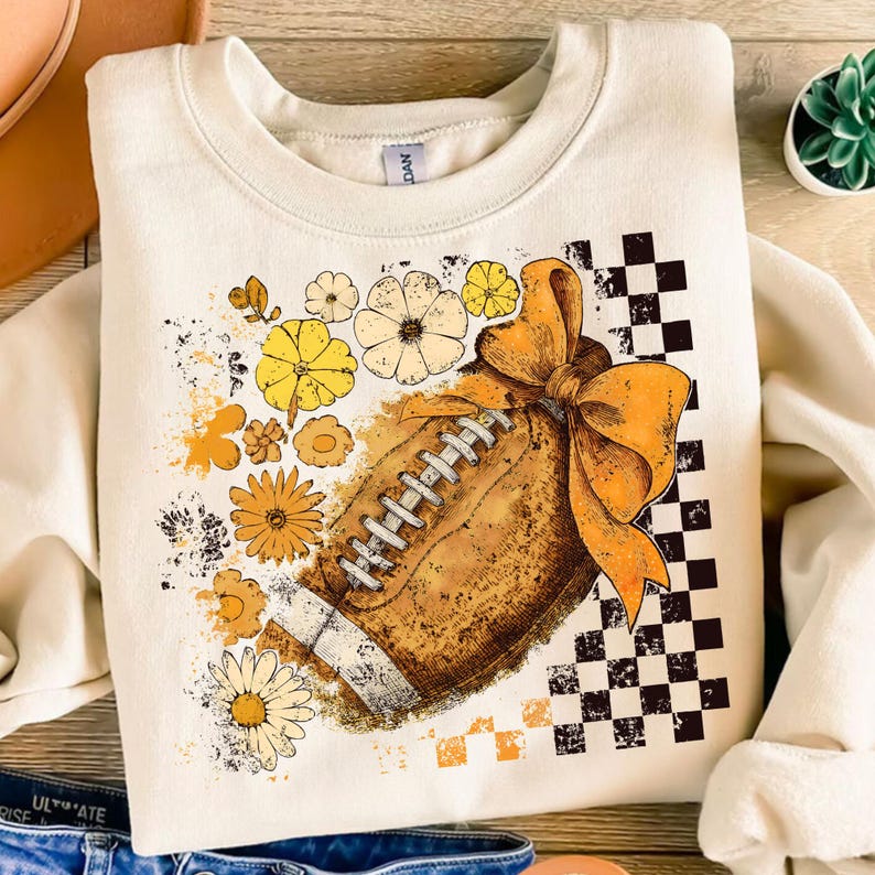 May include: Cream-colored sweatshirt featuring a football design with a large orange bow and floral accents in yellow and white. A black and white checkered pattern is on the right side of the design.
