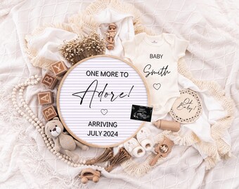 May include: A white baby onesie with the text "Baby Smith" and a small heart. A white and brown wooden sign with the text "One More to Adore! Arriving July 2024" and a small heart. A wooden sign with the text "Oh Baby!" and a small wooden toy.