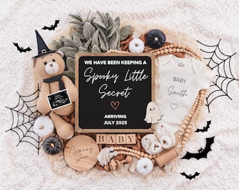 Op de afbeelding: Een Halloween-thema baby aankondiging met een zwart letterbord waarop staat "We have been keeping a spooky little secret. Arriving July 2025." Het letterbord is omringd door een teddybeer met een heksenhoed, een baby onesie met de tekst "Baby Smith", een spinnenweb, vleermuizen en andere Halloween-decoraties.