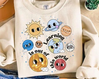 May include: Cream-colored sweatshirt featuring a colorful planet graphic with the text "Learning is out of this world." The design includes a smiling sun, planets, and stars, perfect for space enthusiasts and educators.