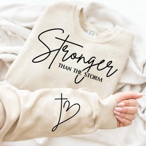 Könnte beinhalten: Beiges Sweatshirt mit dem schwarzen Schriftzug "Stronger Than The Storm". Der Ärmel zeigt ein Kreuz- und Herz-Design. Das Sweatshirt liegt gefaltet auf einer weißen Oberfläche.