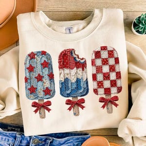 May include: Cream-colored sweatshirt featuring three popsicle designs. The popsicles are decorated with red, white, and blue patterns, including stars and checkered patterns. Each popsicle has a red bow at the base.