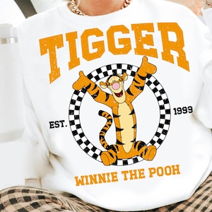 Puede incluir: Sudadera blanca con la palabra "Tigger" en letras naranjas en bloque sobre un personaje de dibujos animados de Tigger. El dise&ntilde;o incluye un c&iacute;rculo a cuadros blanco y negro y el texto "EST. 1999 WINNIE THE POOH".