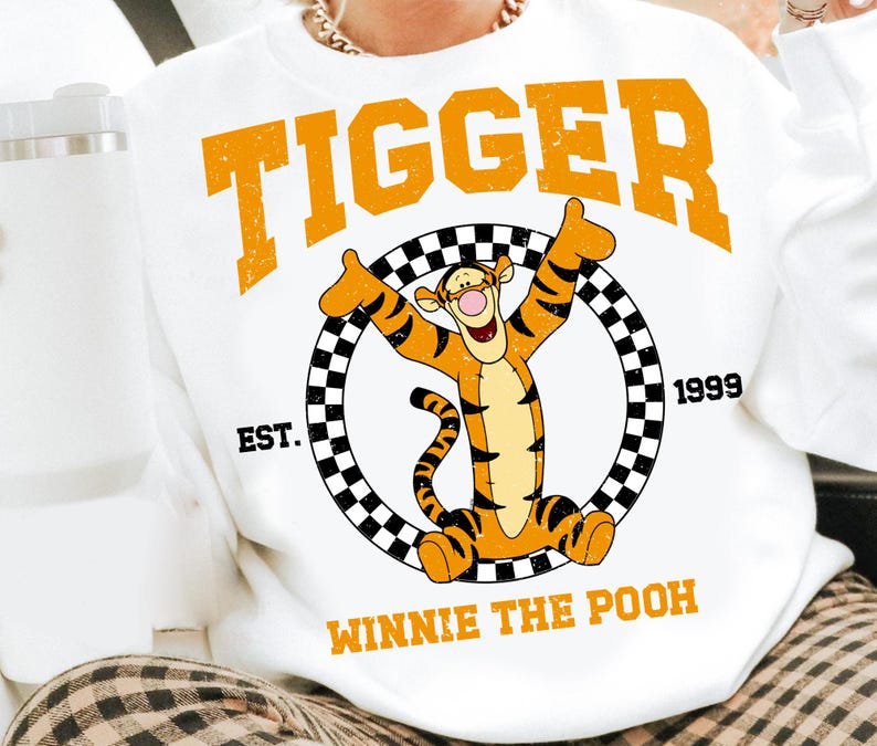 Puede incluir: Sudadera blanca con la palabra "Tigger" en letras naranjas en bloque sobre un personaje de dibujos animados de Tigger. El dise&ntilde;o incluye un c&iacute;rculo a cuadros blanco y negro y el texto "EST. 1999 WINNIE THE POOH".