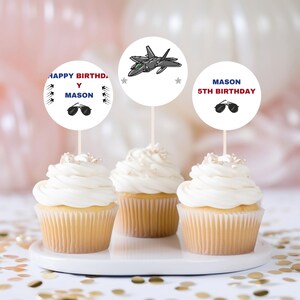 Puede incluir: Tres cupcakes con glaseado blanco y adornos decorativos. Los adornos presentan un avión de combate, gafas de sol y el texto "HAPPY BIRTHDAY MASON" y "MASON 5TH BIRTHDAY". Los cupcakes están sobre un plato blanco con confeti dorado.