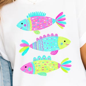 Op de afbeelding: Wit T-shirt met drie kleurrijke visillustraties. Elke vis heeft een uniek ontwerp met roze, blauwe en gele accenten. De vissen hebben lachende gezichten en zijn verticaal op het shirt geplaatst.