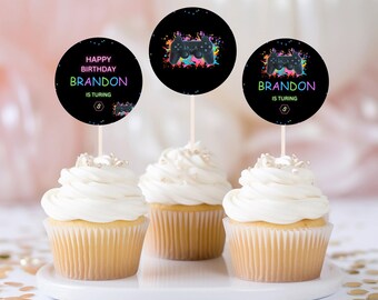 May include: Three cupcakes with white frosting and gold sprinkles topped with round black and neon green, pink, and blue gamer themed cupcake toppers. The toppers feature a video game controller and the text "Happy Birthday Brandon is Turing".