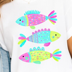 Op de afbeelding: Wit T-shirt met drie kleurrijke cartoonvissen. De vissen hebben roze, blauwe en gele lichamen met verschillende patronen en details. Elke vis heeft een lachend gezicht en een uniek vinontwerp. Een casual kledingstuk.