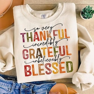以下が含まれることがあります： クリーム色のスウェットシャツ。カラフルなレイヤーテキストで「So Very Thankful, Incredibly Grateful, Unbelievably Blessed」と書かれています。長袖とクルーネックで、秋に最適です。