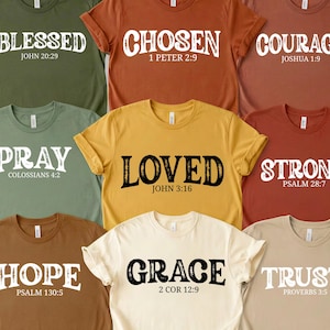 May include: A collection of crew neck t-shirts in various colors, each featuring a different inspirational word in bold white lettering. The words include "Blessed," "Chosen," "Courage," "Pray," "Loved," "Strong," "Hope," "Grace," and "Trust," along with corresponding Bible verses.
