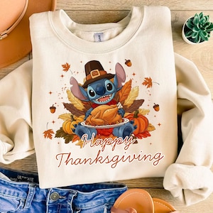 May include: Cream-colored sweatshirt featuring a cartoon character wearing a pilgrim hat and holding a roasted turkey. The graphic includes pumpkins, autumn leaves, and the text "Happy Thanksgiving" in a script font. A festive design.
