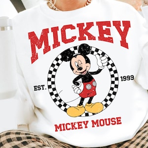 Puede incluir: Sudadera blanca con un gr&aacute;fico de Mickey Mouse haciendo una se&ntilde;al de aprobaci&oacute;n, dentro de un c&iacute;rculo a cuadros. La palabra "MICKEY" est&aacute; impresa en rojo encima del gr&aacute;fico, con "MICKEY MOUSE" debajo. La sudadera tambi&eacute;n tiene impreso "EST. 1999".