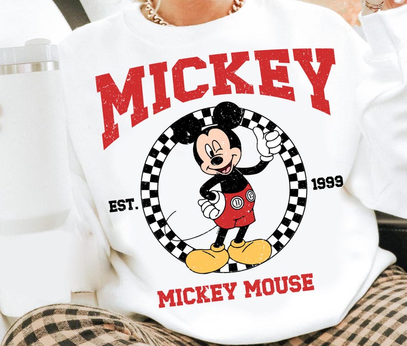 Puede incluir: Sudadera blanca con un gr&aacute;fico de Mickey Mouse haciendo una se&ntilde;al de aprobaci&oacute;n, dentro de un c&iacute;rculo a cuadros. La palabra "MICKEY" est&aacute; impresa en rojo encima del gr&aacute;fico, con "MICKEY MOUSE" debajo. La sudadera tambi&eacute;n tiene impreso "EST. 1999".