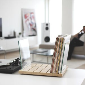 May include: A light-colored wooden vinyl record storage rack featuring two clear glass side panels. The rack holds a collection of vinyl records with various colored sleeves, positioned next to a black turntable on a white surface.