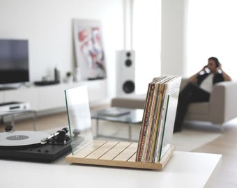 Könnte beinhalten: Ein Schallplattenständer aus hellem Holz mit zwei seitlichen Paneelen aus klarem Glas. Der Ständer enthält eine Sammlung von Schallplatten mit verschiedenfarbigen Hüllen und steht neben einem schwarzen Plattenspieler auf einer weißen Fläche.