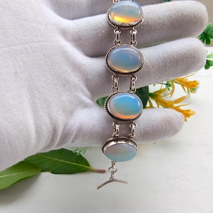 Puede incluir: Una pulsera de plata con cinco piedras cabujón ovaladas y translúcidas con un brillo lechoso e iridiscente. Cada piedra está engastada en un marco de plata trenzada y unida por pequeños eslabones de plata, con un cierre de barra simple.