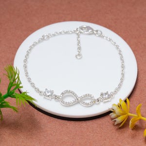Peut inclure: Un bracelet en argent orné d'un symbole infini serti de petits pierres précieuses transparentes. Le bracelet a une chaîne délicate et deux accents de pierres précieuses supplémentaires. Le bijou est présenté sur une surface circulaire blanche sur fond marron.