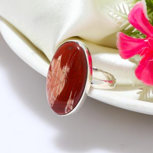 May include: A silver ring featuring a large, oval-shaped gemstone with a deep red and cream-colored pattern. The gemstone is set in a polished silver band. The ring is displayed on a white surface with a pink flower in the background.