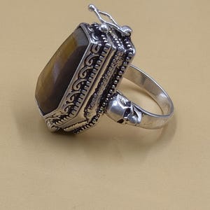 May include: A silver ring with a rectangular, brown gemstone. The ring features ornate detailing around the stone and a skull design on the band. The ring has a unique, gothic aesthetic.